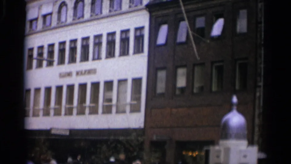 Final 1965 Copenhagen frame with a white lettered facade, adjacent brick building, street-level pedestrians, awnings, and a rounded foreground fixture.