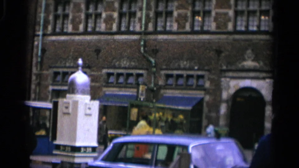 1965 Copenhagen street view with a passing car, pedestrians, dark brick buildings, awnings, and a white domed street structure.