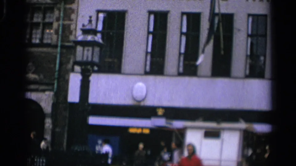 1965 Copenhagen street view with a pale multi-window facade, dark awnings, a street lamp, and people moving along the sidewalk.