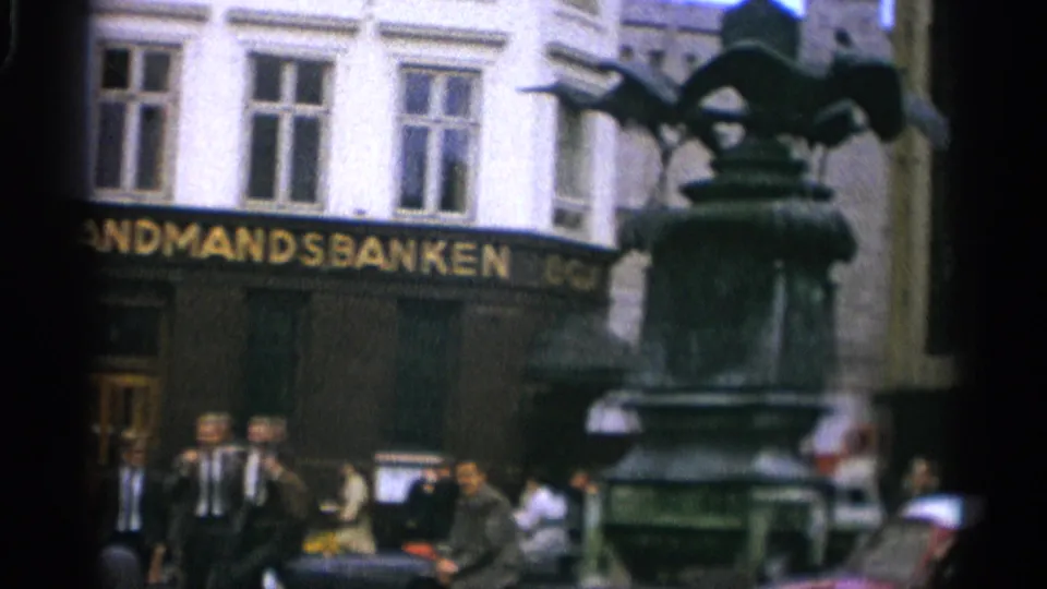 A bronze statue or fountain base partially blocks the Landmandsbanken facade as pedestrians stand and walk nearby in Copenhagen.