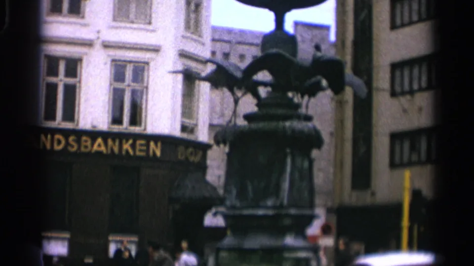 1965 Copenhagen frame with the fountain shifted to the right, partial bank lettering visible behind it, pedestrians near the entrance, and urban facades under overcast light.