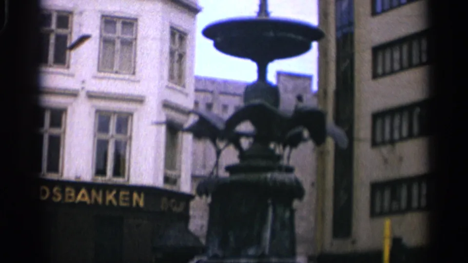 1965 Copenhagen frame emphasizing the upper fountain bowl and sculpted birds, with pale building windows, partial bank signage, and a narrow street view behind it.
