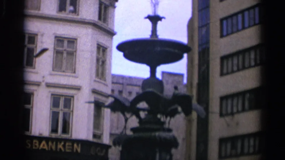 Final 1965 Copenhagen frame showing the fountain silhouetted against cloudy sky and pale facades, with partial bank signage and adjacent street architecture visible behind it.
