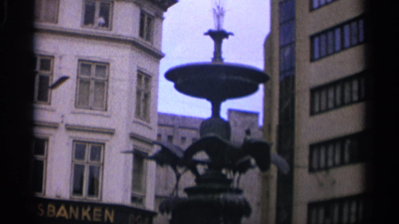 1965 Copenhagen street view with a dark decorative fountain framed by multi-story buildings and partial bank signage.