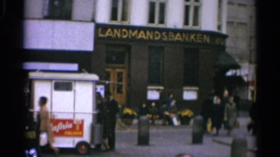 A white ice cream cart with red branding stands before Landmandsbanken as pedestrians pass through the square.