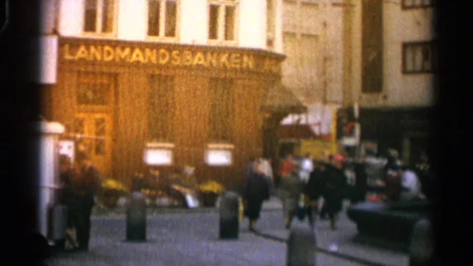 Warm exposed 8mm frame of pedestrians moving past Landmandsbanken, with bollards and a partial fountain edge visible.