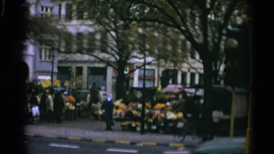 1965 Copenhagen street corner with pedestrians, a flower stall, trees, and building facades seen in soft 8mm color.