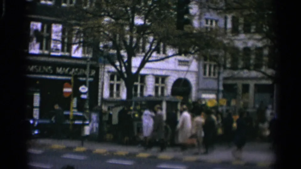 1965 Copenhagen pedestrians cross in front of storefronts and a prominent arched building entrance near the flower market.