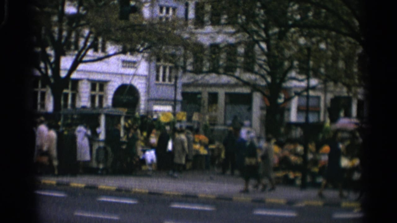 1965 Copenhagen street-market view with pedestrians, produce stands, trees, and urban building facades seen from across the road.