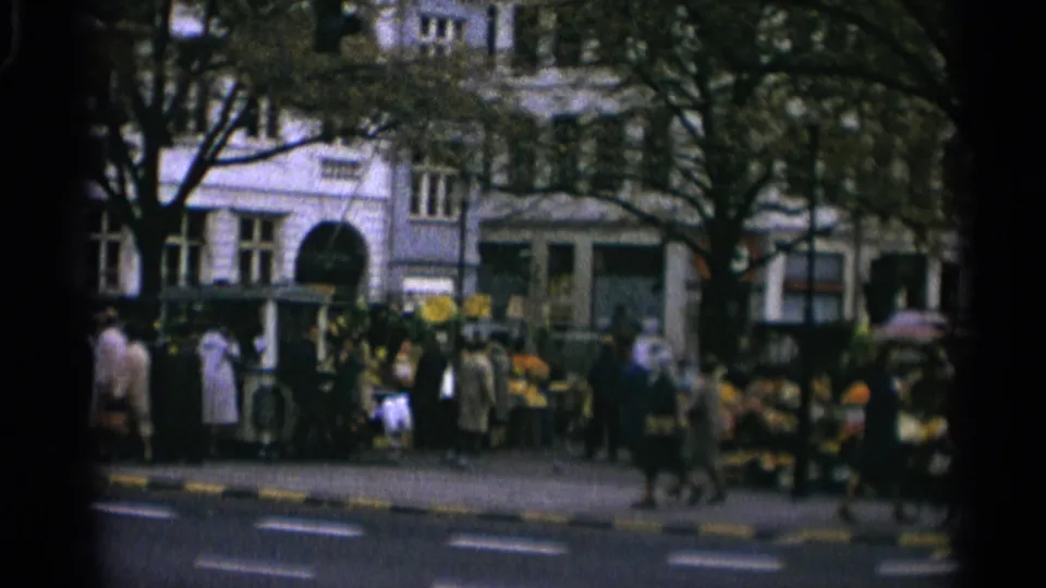 1965 Copenhagen street-market view with pedestrians, produce stands, trees, and urban building facades seen from across the road.