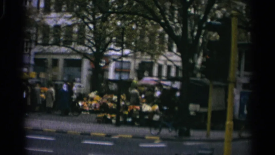 Copenhagen market scene with pedestrians near produce stands, a bicycle at the curb, a yellow street pole, trees, and apartment-style facades.