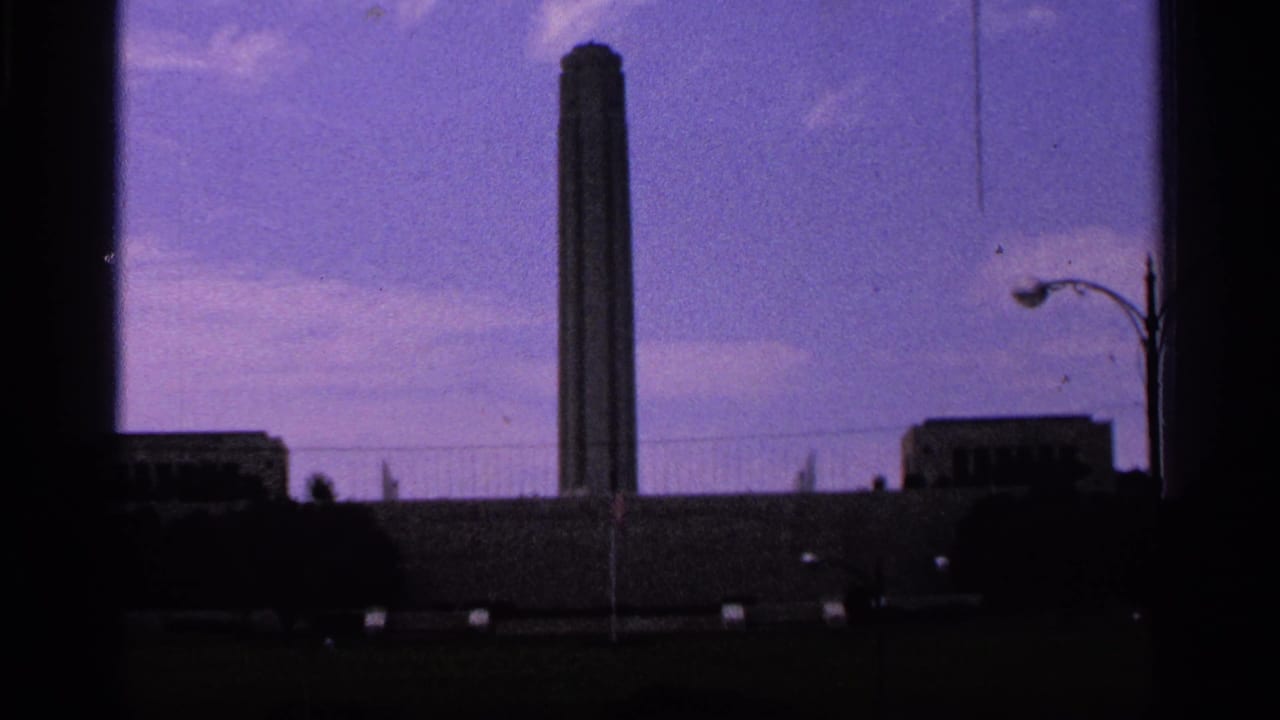 1965 Kansas City home-movie frame showing the Paseo Arch area against a purple sky, with dark frame edges and a streetlight at right.