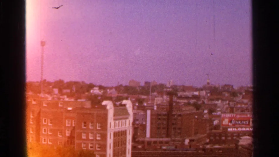 Elevated 1965 Kansas City view across brick buildings and distant structures, with a visible rooftop business sign and warm flare from the 8mm film scan.