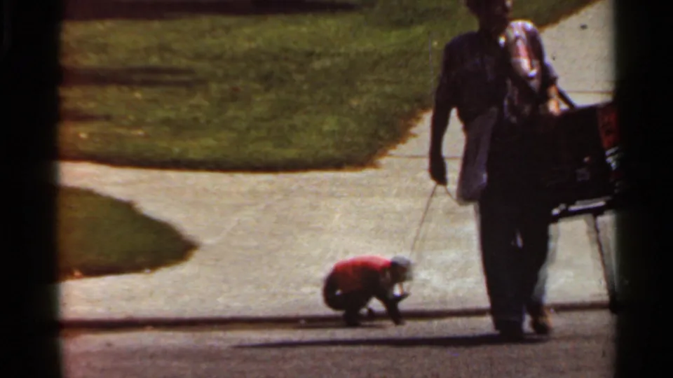 A man with a carried box walks past the leashed monkey in red on a sunlit sidewalk in Kansas City.