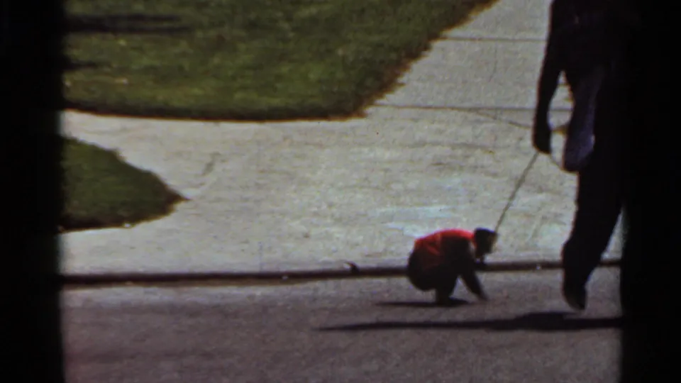The small monkey in a red garment crouches near the curb while the handler exits the right side of the frame.