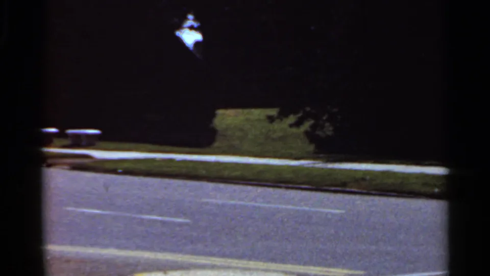 1965 Kansas City home-movie frame showing a dim street, lane markings, curbside grass, and trees in low light.