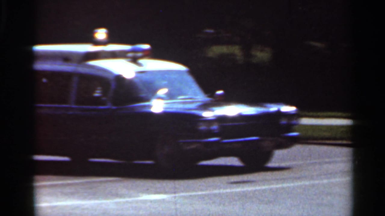 Vintage police car passes on a dark road with a roof light visible.