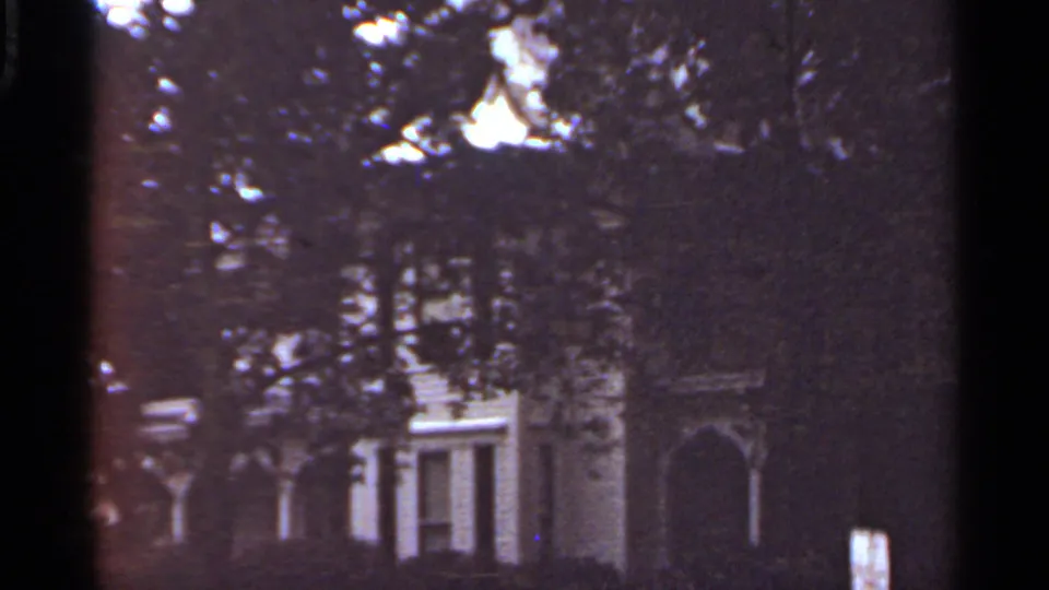 A white house exterior is partly hidden by trees in a dark 1965 Kansas City residential home-movie frame.