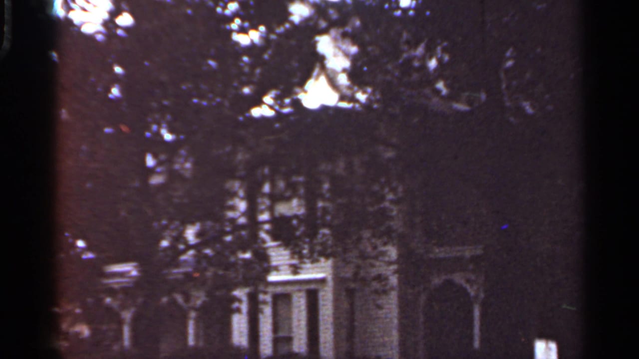 1965 Kansas City 8mm frame showing a house exterior partly hidden behind dense tree foliage and dark shadows.