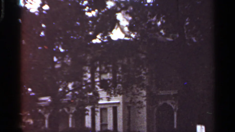 1965 Kansas City 8mm frame showing a house exterior partly hidden behind dense tree foliage and dark shadows.