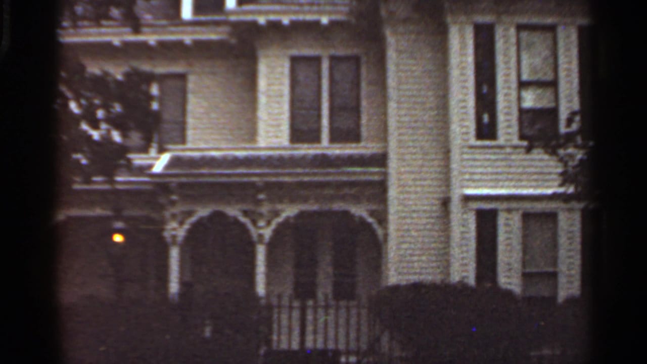 Dim view of a light-colored multi-story house with porch arches and trees.