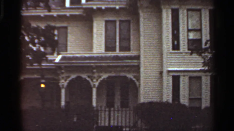 1965 Kansas City home-movie frame showing a light-colored multi-story house with ornate porch arches, trees, and a glowing exterior light.