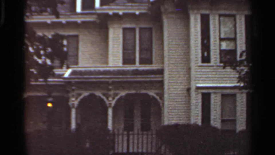 1965 Kansas City frame of a light-sided residential facade with ornate porch arches, dark windows, tree shadow, and a small exterior light.
