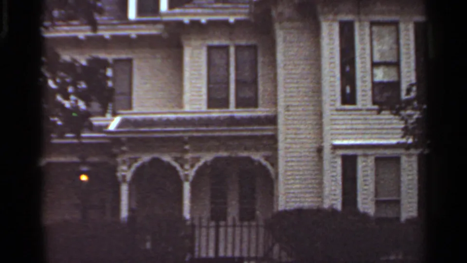 1965 Kansas City home-movie view of a Victorian-style house exterior with porch arches, railing, shrubs, and a small glowing exterior light.