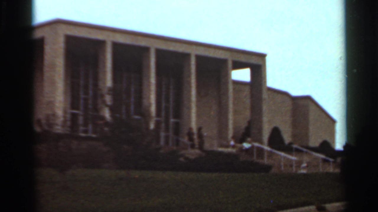 Large modernist building with columns, glass bays, lawn, steps, and distant people near the entrance.