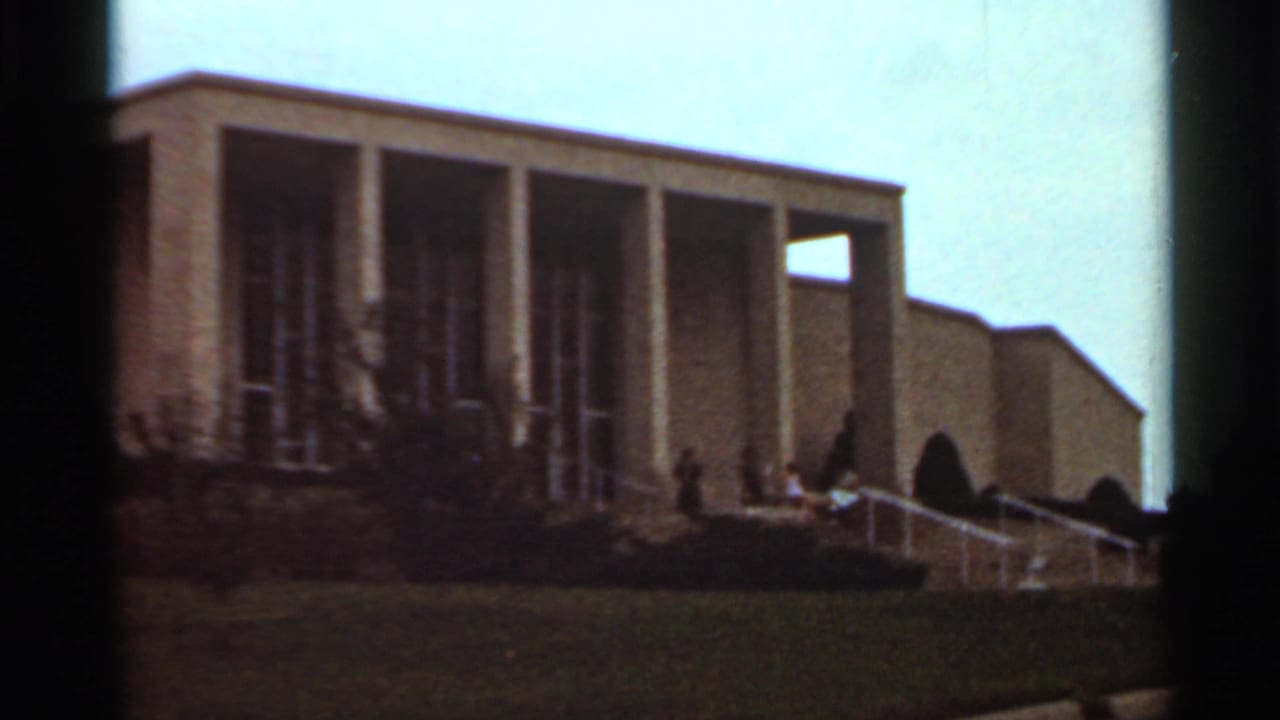 Columned building facade with lawn, steps, railings, shrubs, and small distant figures near the entrance.