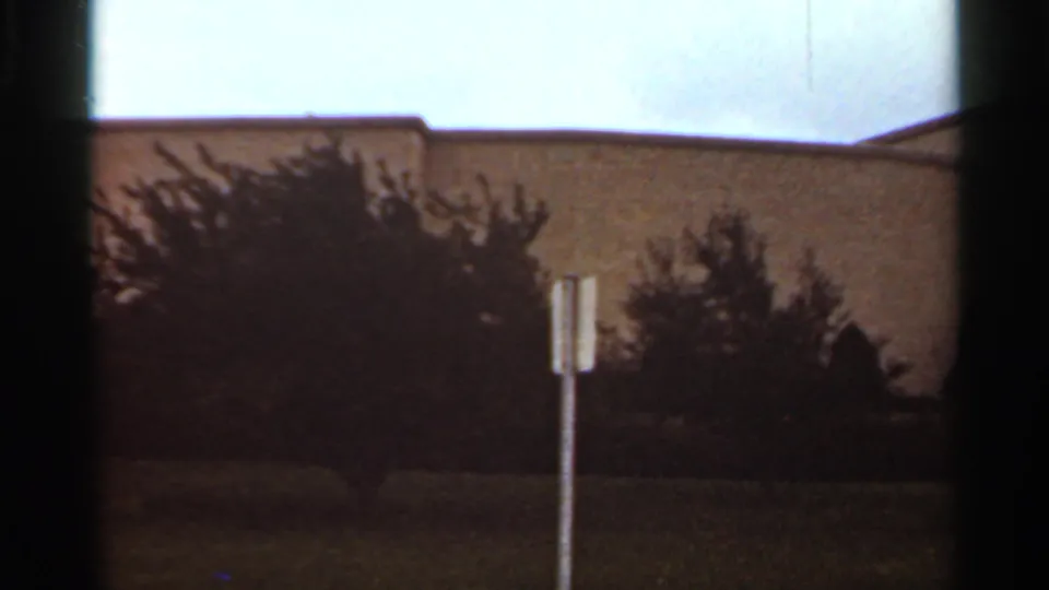 1965 Kansas City footage shifts to a side or nearby wall of the large building, with shrubs, a small signpost, and grass in the foreground.