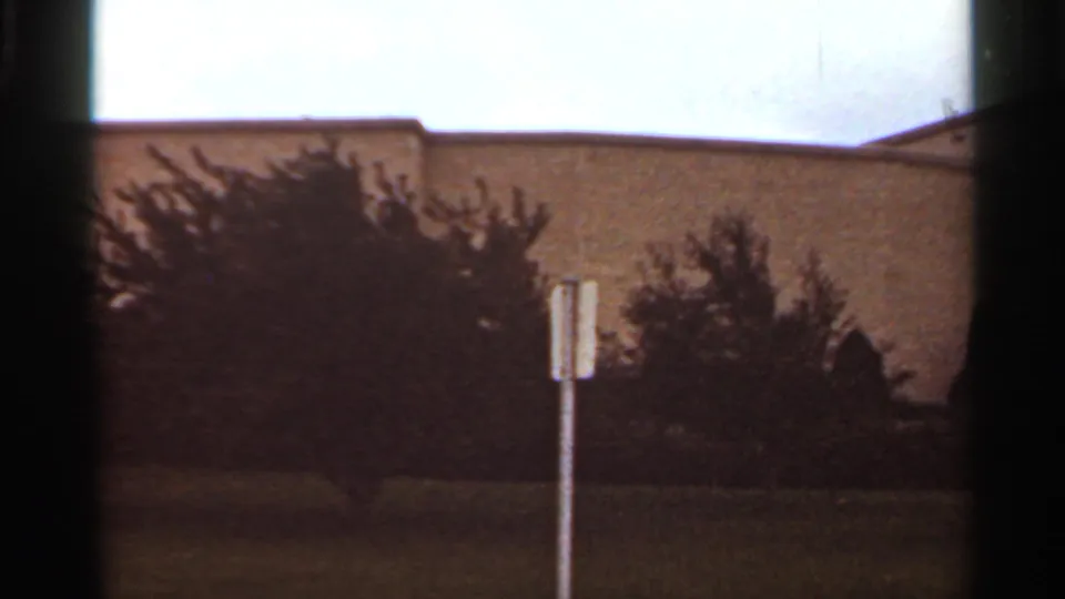 Kansas City, 1965, showing shrubs and a small signpost in front of a light-colored building wall, filmed with soft 8mm color and dark frame edges.