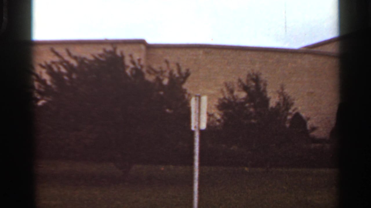 1965 Kansas City 8mm frame showing grass, silhouetted bushes, a pale street sign, and a large plain building under an overcast sky.