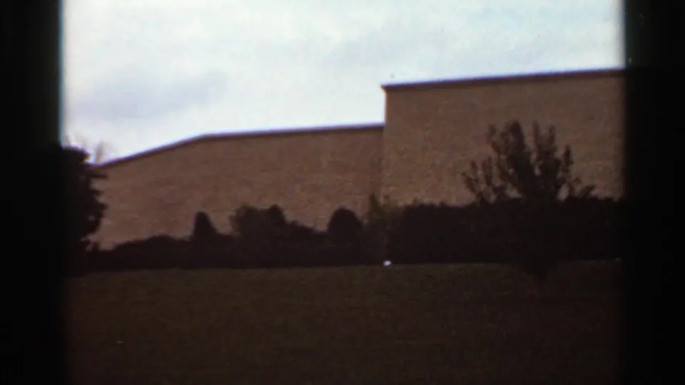 Kansas City 1965 home-movie frame showing grass, dark bushes, and a pale low building with a simple roofline under cloudy light.