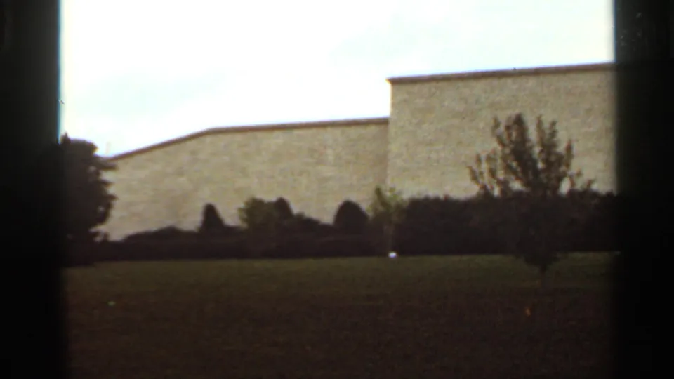 1965 Kansas City 8mm frame with open grass, low dark shrubs, a pale flat-roofed building, and muted overcast light.
