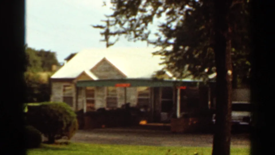 1965 home-movie frame showing the Cortez Motel building exterior with a white roof, green trim, trees, and a parked vehicle.