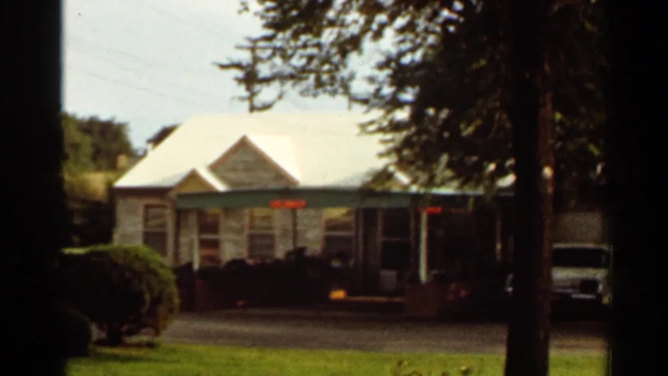 Kansas City area motel exterior from a 1965 home movie, with white roof, shaded trees, lawn, driveway, and parked vehicle.