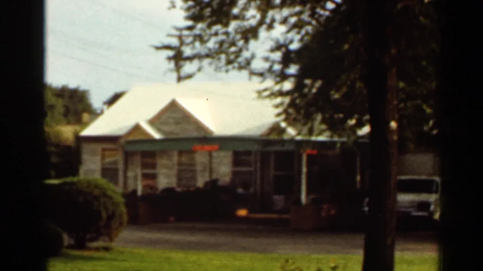 1965 Kansas City home-movie frame showing a stone residential house, lawn, shrubs, and tree-framed driveway view.