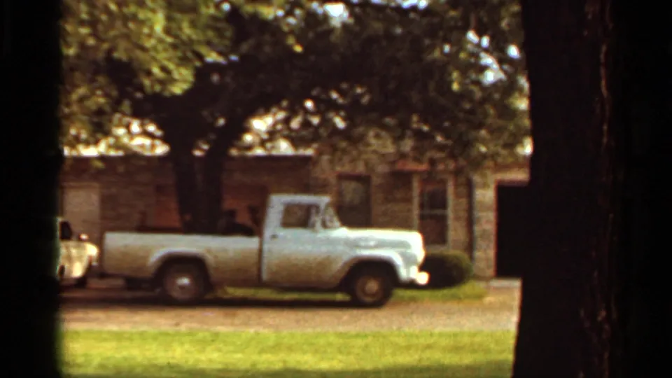 1965 Kansas City frame with a light-colored midcentury pickup parked near a stone house and mature trees.