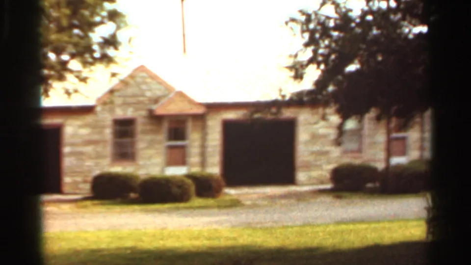 1965 Kansas City home-movie frame showing a stone house facade, dark garage door, shrubs, lawn, and driveway.