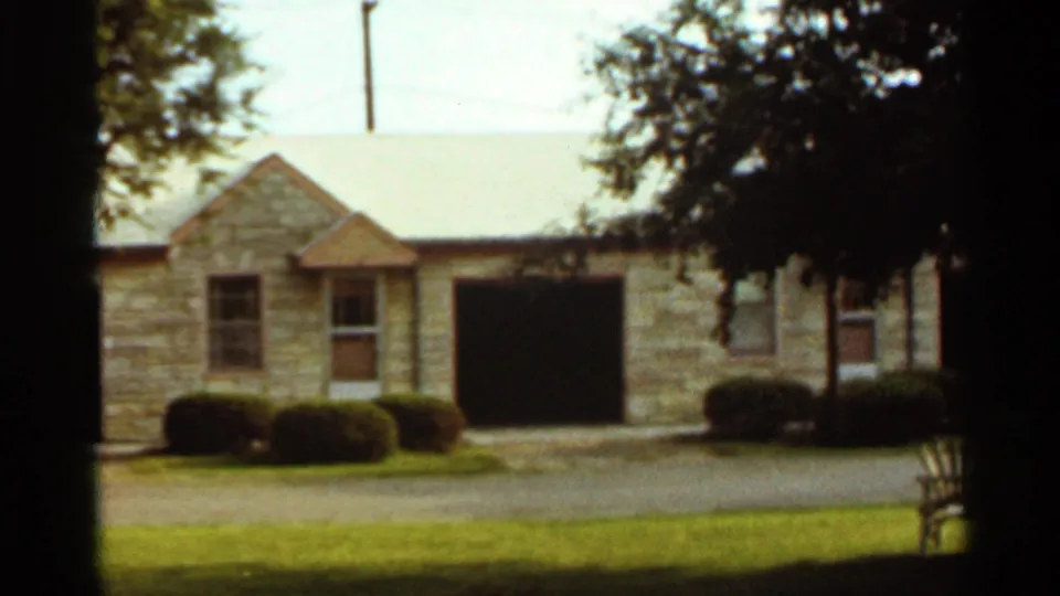 1965 Kansas City frame of a stone residential exterior with garage, lawn, shrubs, and tree shade.