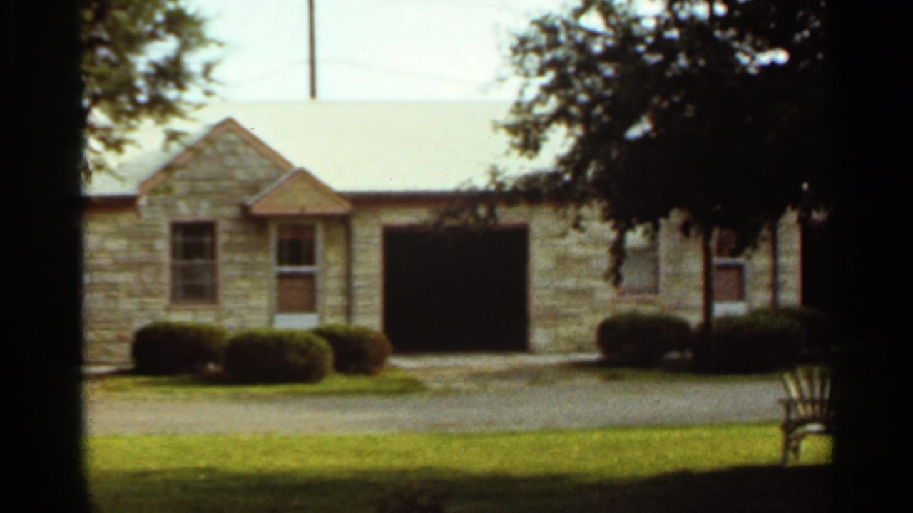 1965 Kansas City suburban house exterior with stone facade, open garage bay, manicured shrubs, lawn, and trees.
