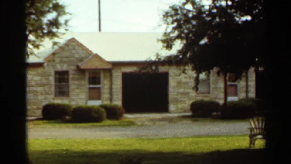 1965 Kansas City suburban house exterior with stone facade, open garage bay, manicured shrubs, lawn, and trees.