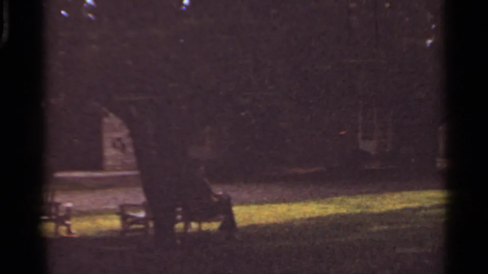 Dim 1965 Kansas City yard view with two people seated on a bench beneath large trees near a house exterior.