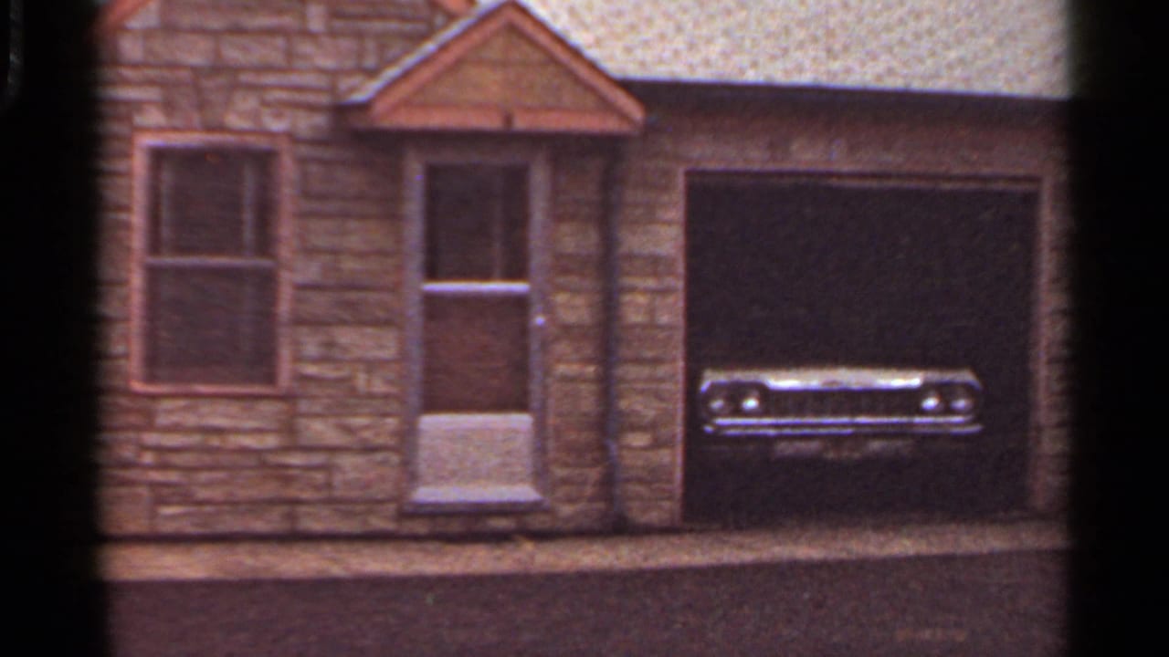1965 8mm frame showing a stone-faced house exterior with a garage and the front of a parked mid-century car.