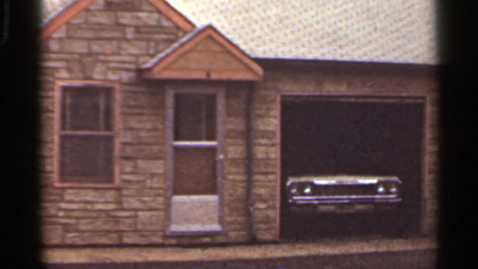 1965 home-movie view of a stone residential exterior, garage bay, and a car parked inside the opening.