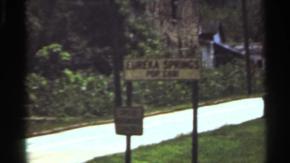 1965 8mm travel frame showing a Eureka Springs population sign beside a road and wooded roadside.