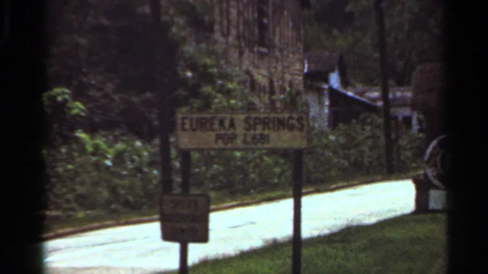 1965 home-movie frame with a roadside Eureka Springs sign and population placard along a wooded road.