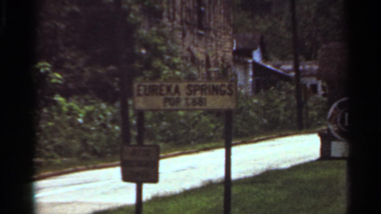 1965 8mm frame showing a Eureka Springs population sign beside a quiet road and wooded roadside.