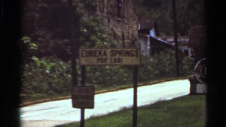 1965 8mm frame showing a Eureka Springs population sign beside a quiet road and wooded roadside.